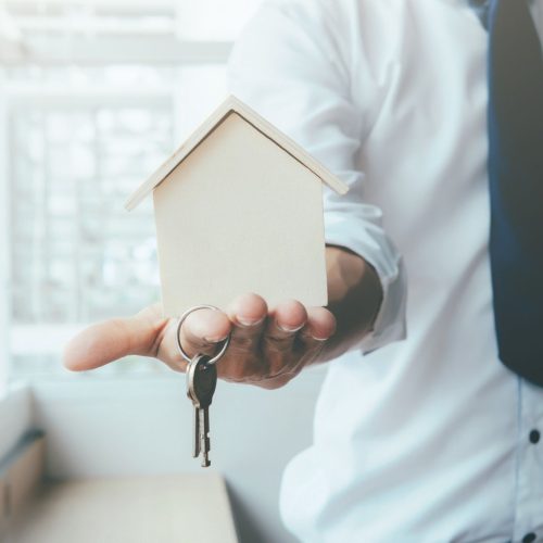 Hand agent with home in palm and key on finger. Offer of purchase house, rental of Real Estate. Giving, offering, demonstration, handing house keys.